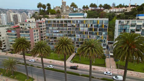 dolly out fly over a residential building in vina del mar, with palm trees and brunet castle in the background, cloudy day and high traffic, chile.