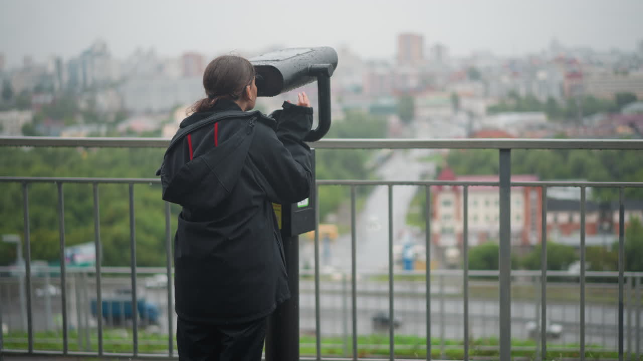 Woman in rain at viewpoint, using public binoculars on balcony, wearing dark raincoat, overcast skyline blurred by drizzle, puddles glistening on wet railing, distant traffic and buildings softened