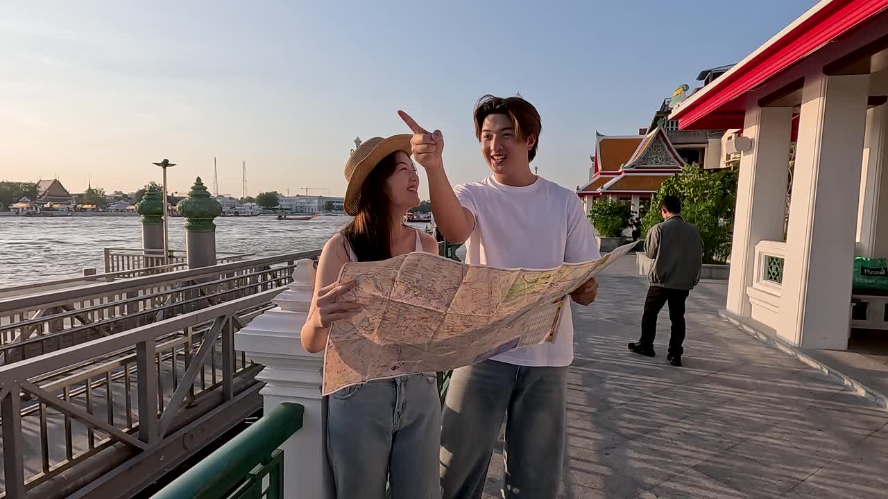 Smiling young tourists consult paper map on riverside promenade near temple during warm sunset