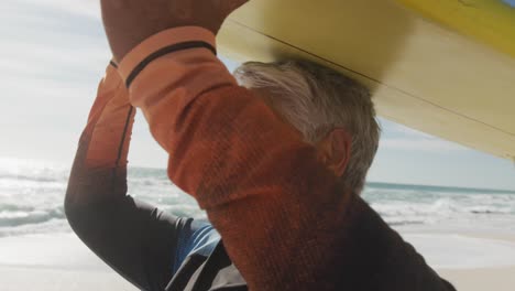 Profile-of-happy-senior-hispanic-woman-walking-on-beach-with-surfboard-over-head