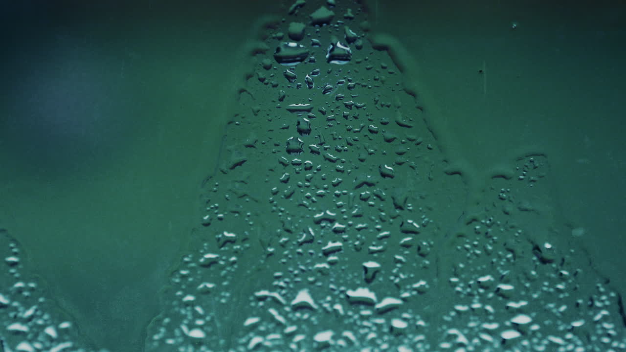 A macro shot of water droplets running down a glass surface, forming patterns and streaks in a muted green color palette