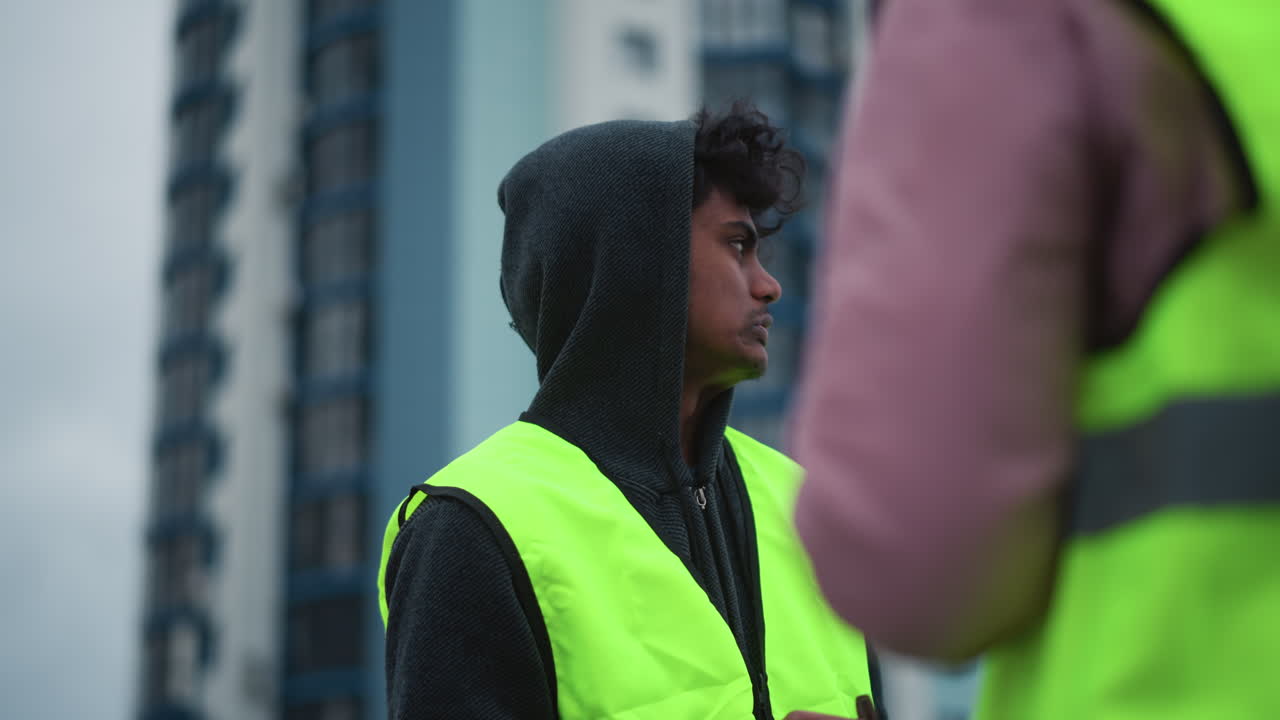 Man in dark hoodie and bright reflective safety vest standing outdoors on cloudy day, looking attentively toward colleague holding sandwich, water bottle visible in background, during cold weather