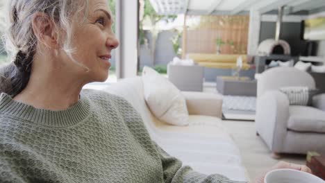 Happy-senior-caucasian-woman-sitting-on-sofa-and-drinking-tea,-slow-motion