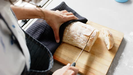 biracial woman wearing apron and cutting fresh bread in kitchen, slow motion