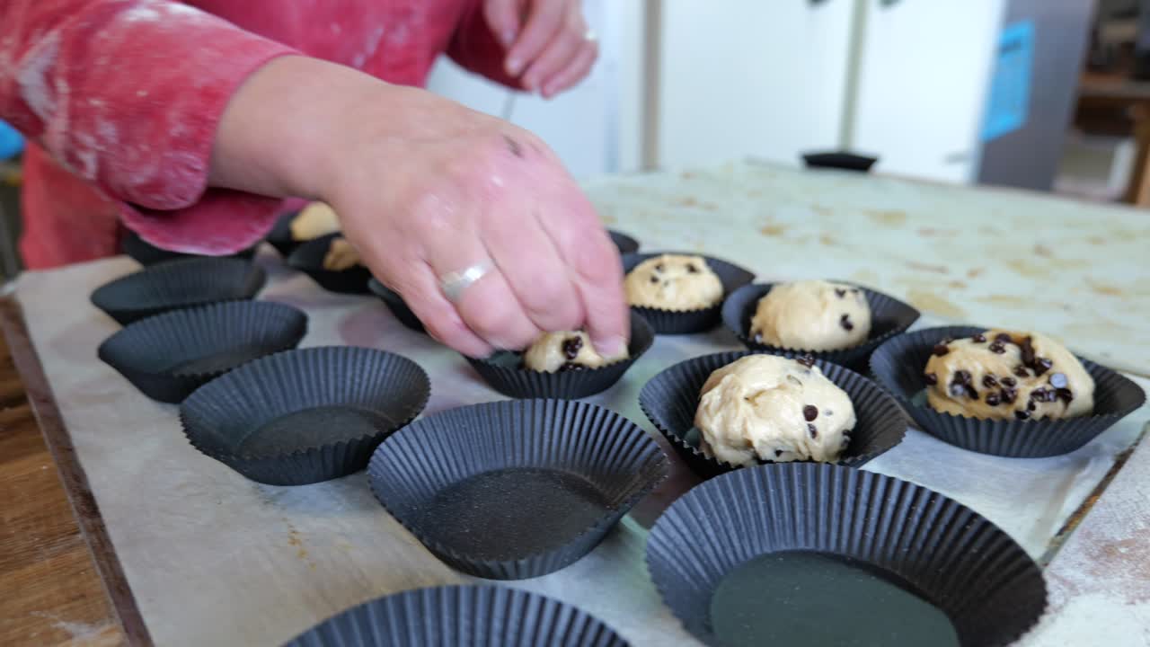 Vista laterale di due fornai che modellano piccoli pezzi di impasto con gocce di cioccolato e li posizionano in singoli stampi da forno su un tavolo di legno infarinato all'interno di un panificio tradizionale