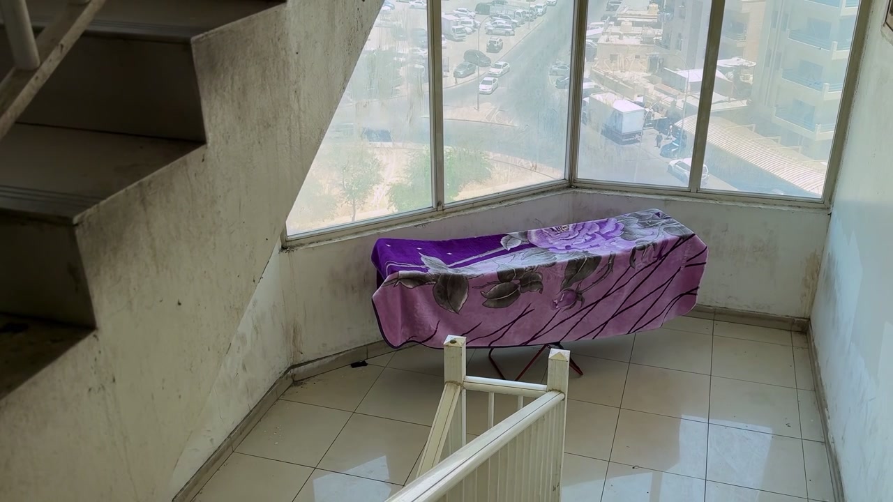 A purple floral blanket dries on a folding rack in a sunlit apartment stairwell landing, with large windows overlooking an urban residential street.