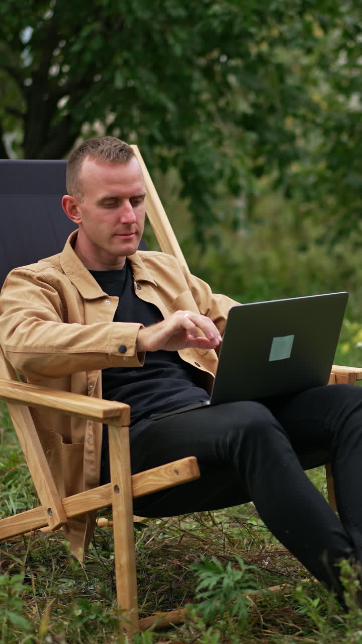 Caucasian male freelancer starting work on his computer. Man sits in a garden chair opening his laptop to start remote work. Nature backdrop. Vertical video