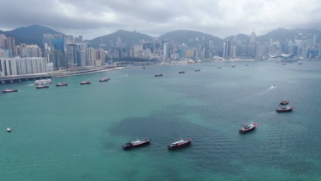 Convoy-De-Barcos-De-Pesca-Locales-Que-Causan-En-La-Bahía-Victoria-De-Hong-Kong,-Con-El-Horizonte-De-La-Ciudad-En-El-Horizonte,-Vista-Aérea