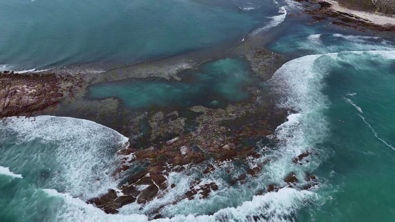 Toma aérea de establecimiento de olas blancas y espumosas y rocas escarpadas a lo largo de la costa de Salmon Hole, piscinas naturales de Daly Head