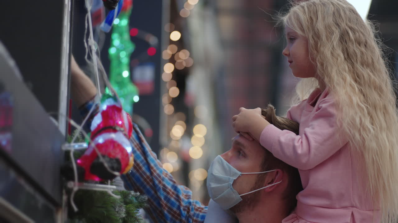 아버지와 딸은 쇼핑몰에서 어깨에 앉아 집에 꽃걸이와 팬데 ⁇  기간 동안 크리스마스에 크리스마스 트리를 슬로우 모션으로 선택합니다.