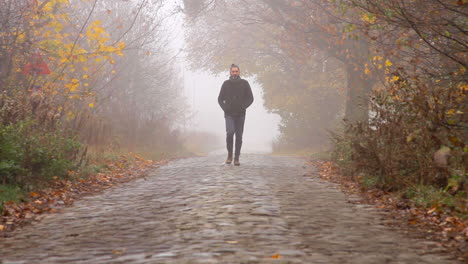 a man is walking towards the camera on a stone paved road during a foggy misty morning