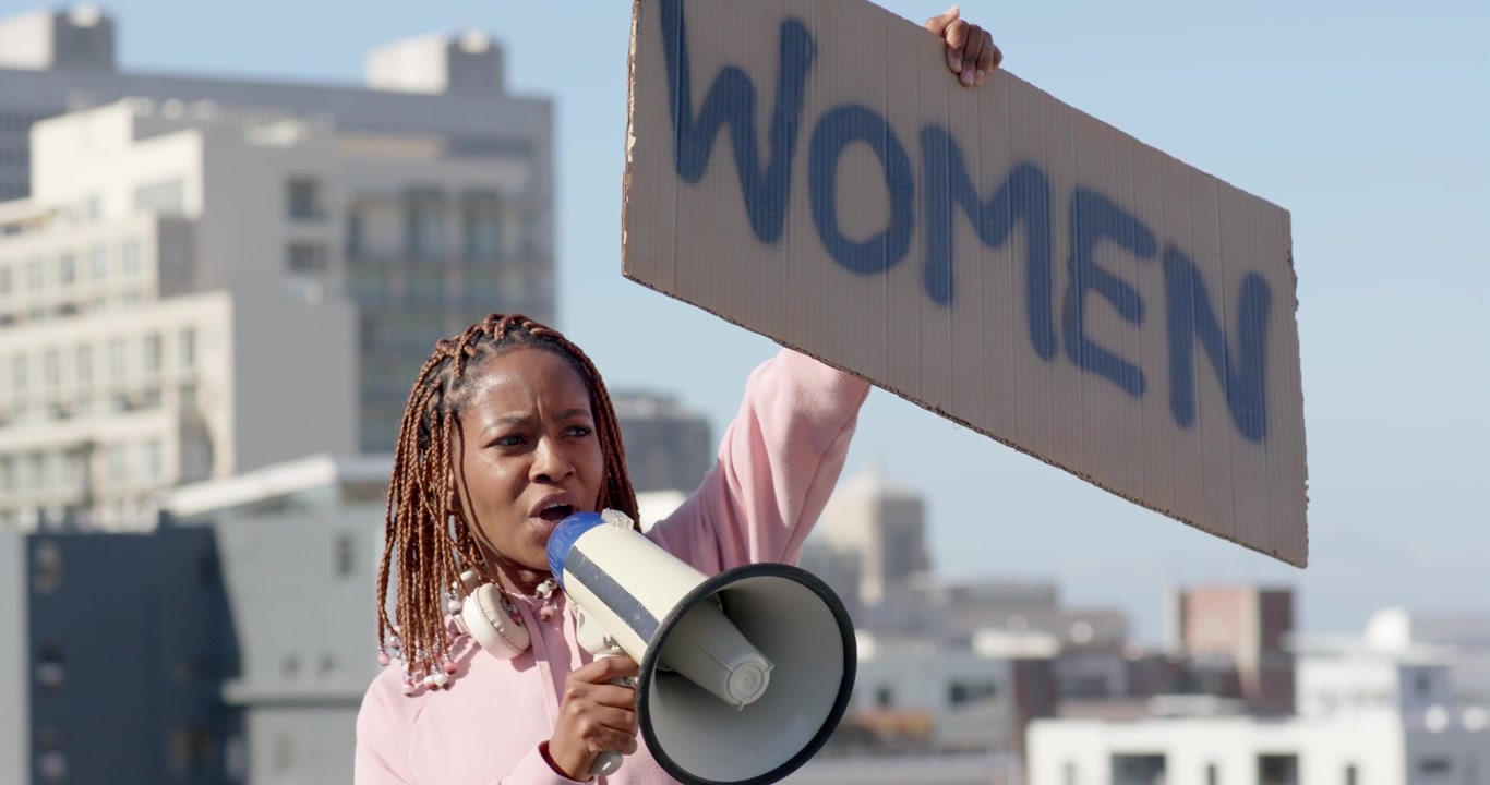 Frau auf Dach hält 'WOMEN'-Schild und spricht in Megafon für Rechte, während einer Kundgebung.
