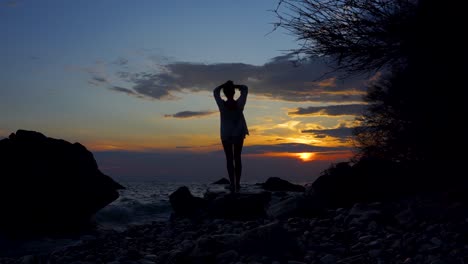 Young-woman-relaxing-on-rocky-shore,-watching-sea-under-colorful-sky-at-sunset-in-Mediterranean