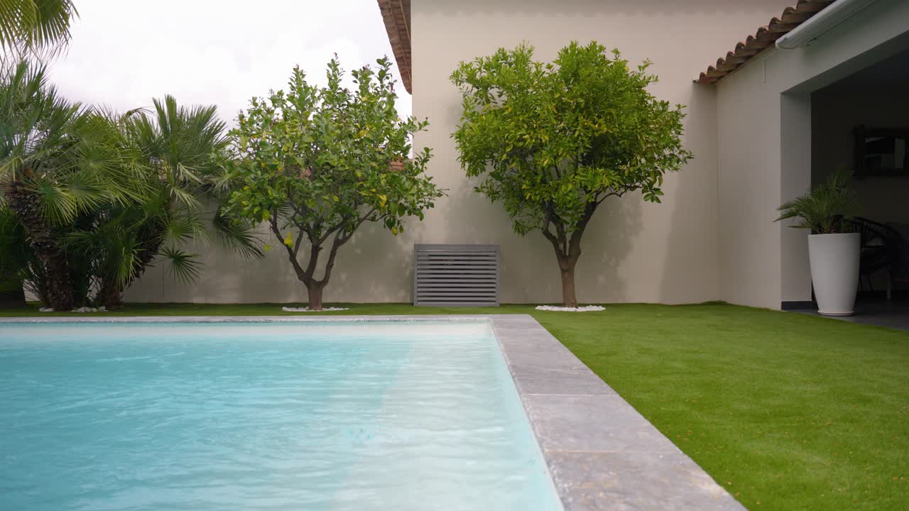 A stylish backyard scene featuring a serene swimming pool, artificial grass lawn, and decorative citrus trees. An exterior AC unit is discreetly hidden with a sleek ventilation cover.