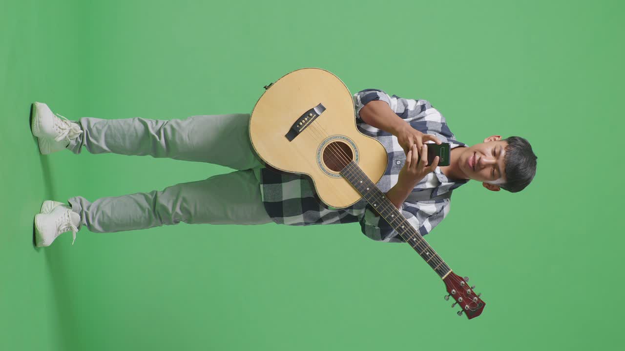 Adolescente asiatico a figura intera con la chitarra che usa lo smartphone mentre è in piedi su sfondo a schermo verde