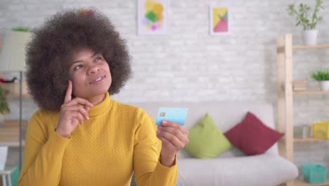 portrait positive and enthusiastic beautiful african american woman with an afro hairstyle looking at the bank card in their hands in the modern apartment