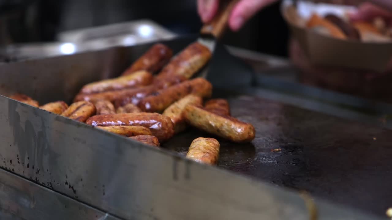 Medium shot of hands flipping vegan sausages on hot flat gas grill with spatula at festival tent. In background, other hand holds hotdog with sausage. Authentic street food cooking atmosphere