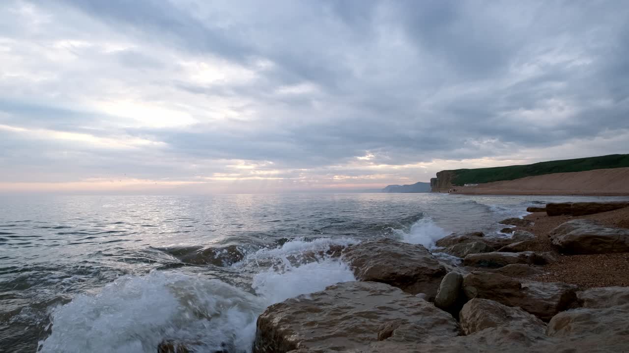 Static low shot of waves crashing against rocks of a Jurassic Coast Beach in Dorset. Captured just after the sun sets behind s the clouds and a grass top cliff is across the bay.