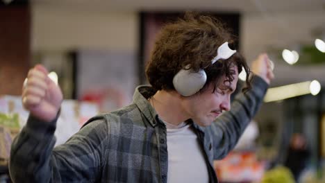 close up of a happy brunette guy with curly hair wearing white headphones dancing and listening to music during his shopping in the supermarket