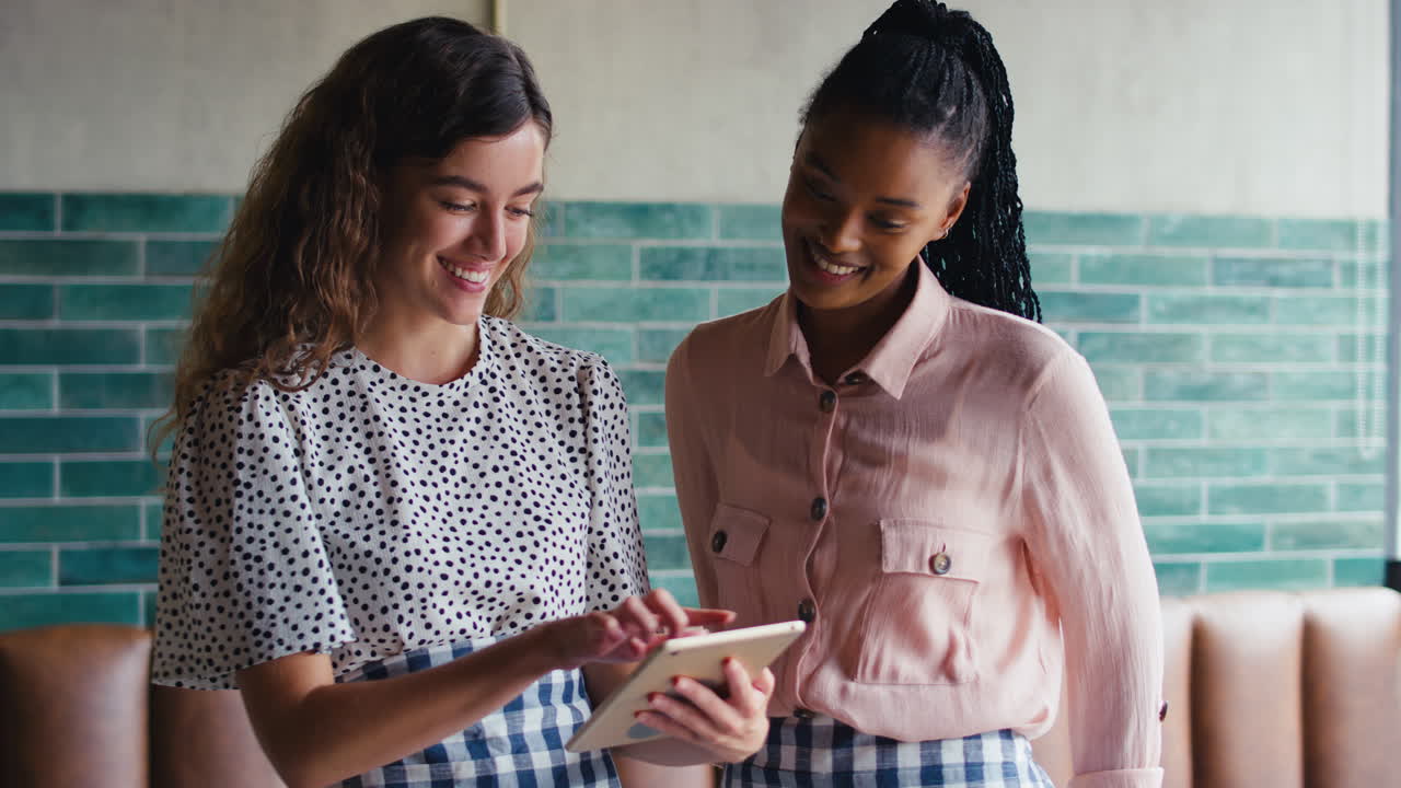 Two Smiling Female Owners Or Staff Looking At Digital Tablet In Cafe Or Coffee Shop