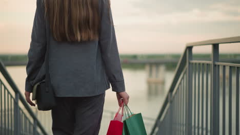 back view of lady walking down stairs carrying colorful shopping bags, with iron railings on both sides and a black carpet underfoot, blurred background features riverfront