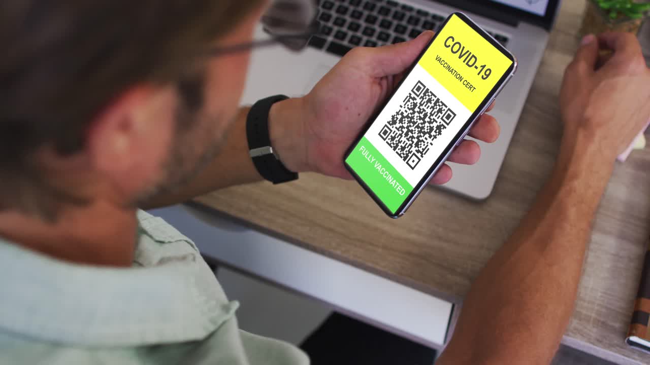 Man at desk holding smartphone with covid vaccination certificate and qr code on screen
