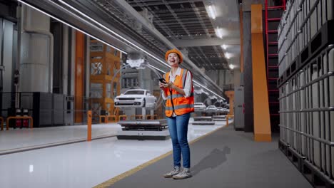 vista lateral completa del cuerpo de una ingeniera asiática con casco de seguridad usando un teléfono inteligente y mirando a su alrededor mientras está de pie en una fábrica de automóviles ev