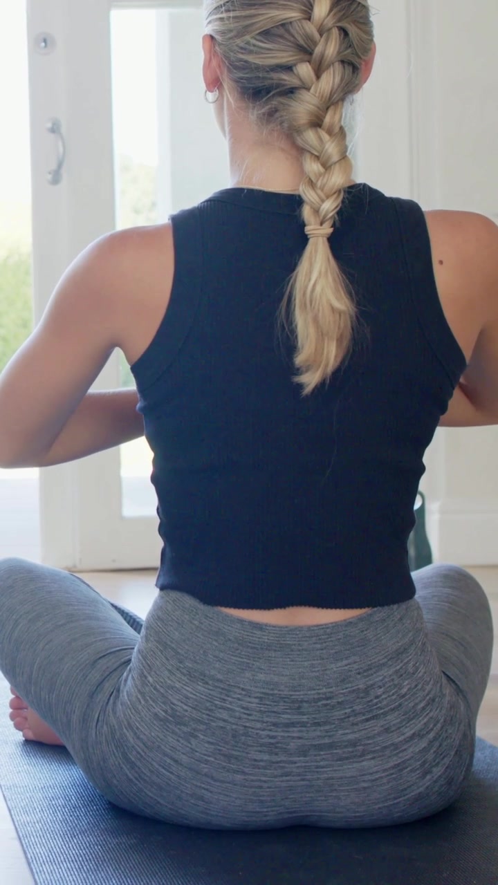 Vídeo vertical: Começando a abaixar, mulher com trança e relógio praticando yoga de top em um tapete perto de portas de vidro