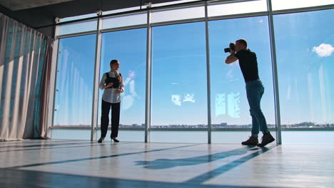 attractive business woman posing to a young photographer. professional male camerist taking photos of a beautiful model in office. panoramic windows.