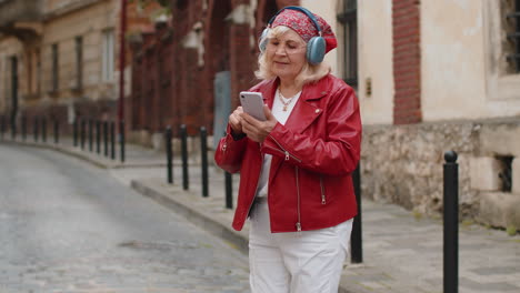 happy senior woman in wireless headphones choosing, listening music in smartphone dancing outdoors