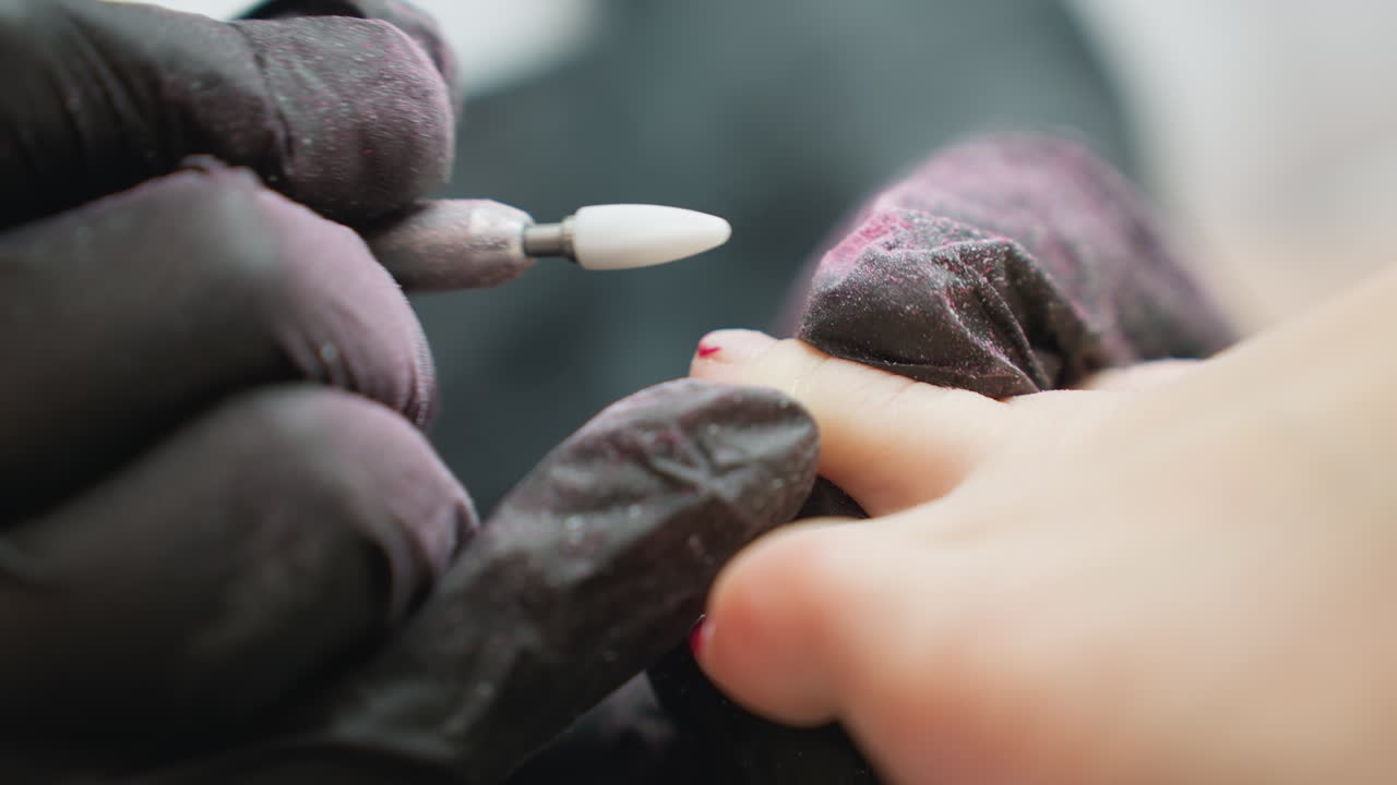 Close up view of nail technician wearing black glove filing client fingernail with electric file, red particles scattered on glove surface, capturing detail of precision work