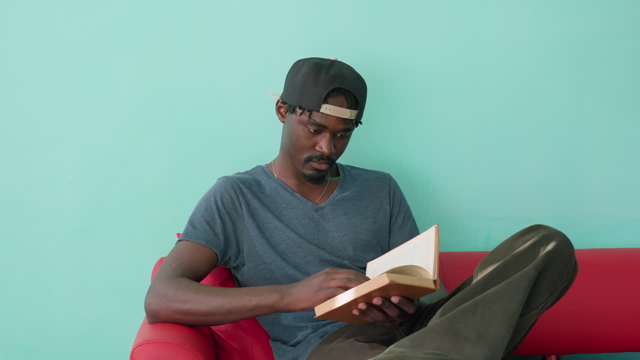 Ein junger Mann in Freizeitkleidung sitzt auf einem roten Sofa, hält ein Buch in den Händen und blättert darin vor einer blaugrünen Wand. Er wirkt nachdenklich und vertieft in einen ruhigen Lesemoment in Innenräumen.