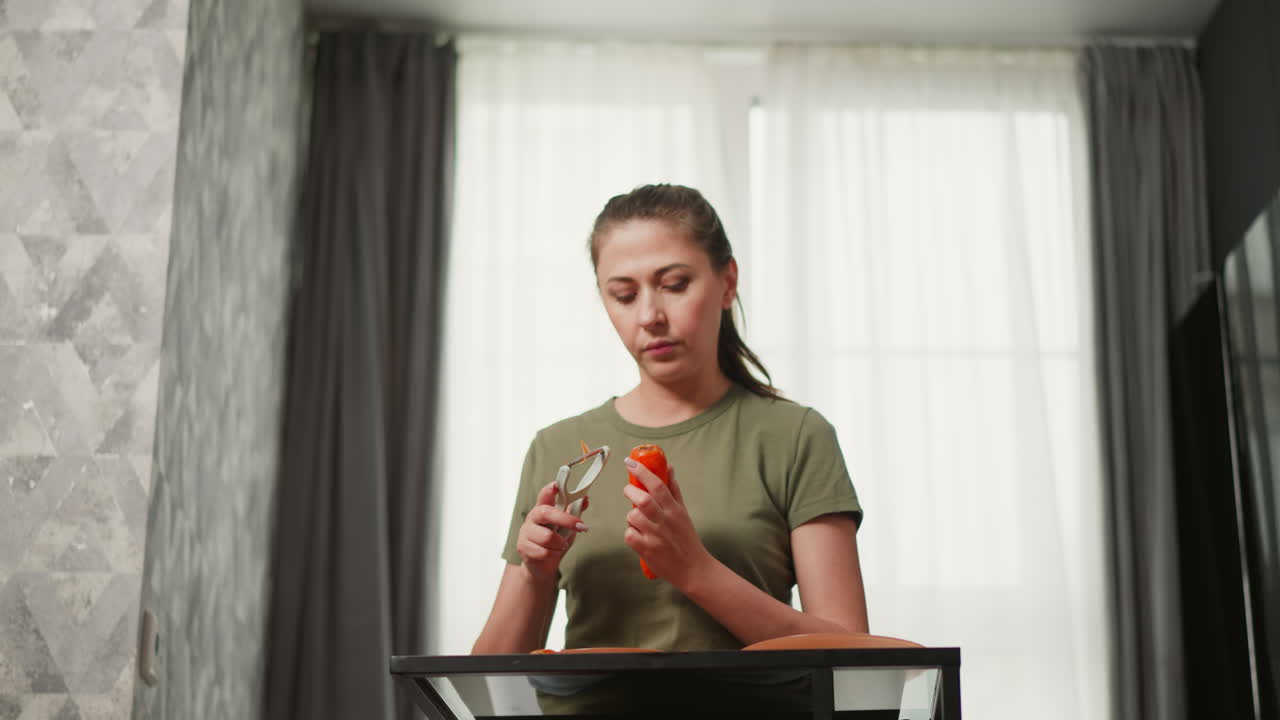 Focused woman in green shirt peeling carrot with handheld peeler while standing at black table, natural daylight streaming through sheer curtains in cozy room with patterned wall background