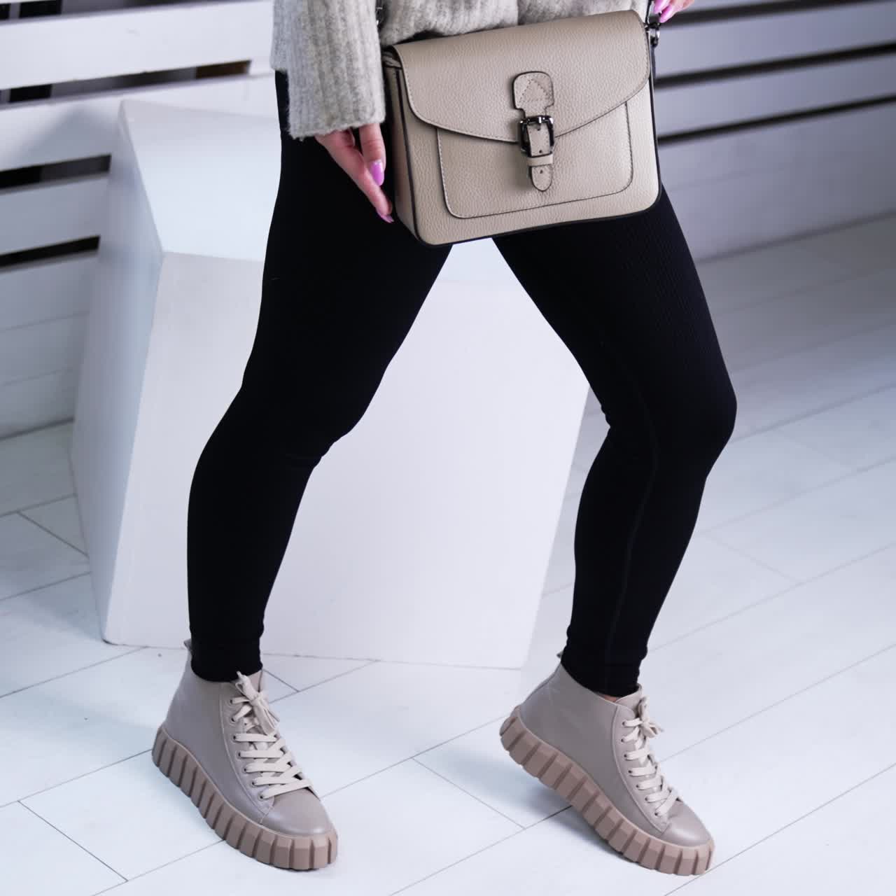 Model in warm sweater and black pants holding a grey bag. Female demonstrating fashionable shoes indoors. White backdrop