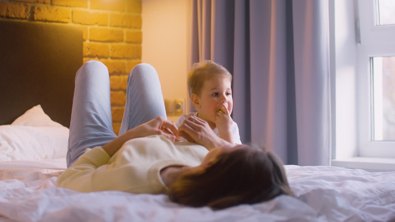 그녀의 아기가 침실에서 장난감을 물고 앉아있는 동안 침대에 누워있는 여자