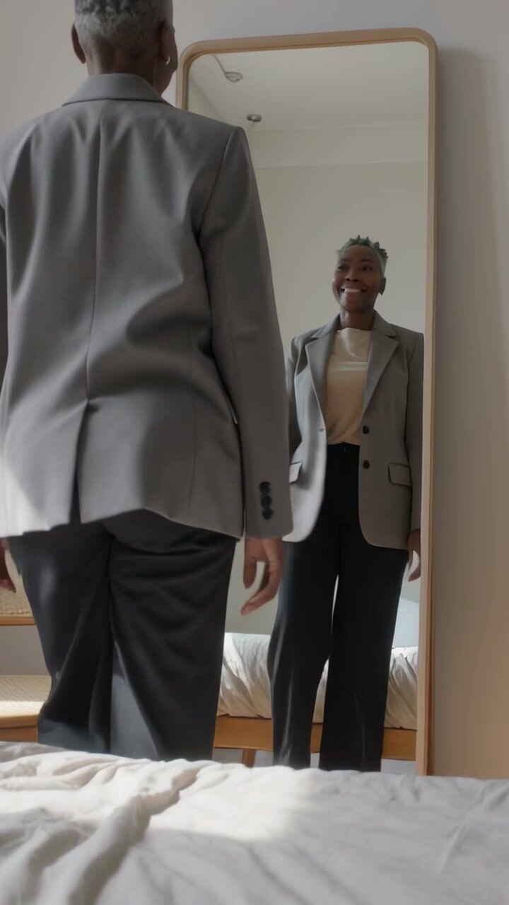 Video verticale: Donna sorridente in blazer che si sposta dal letto allo specchio a casa, controllando l'outfit per il lavoro.
