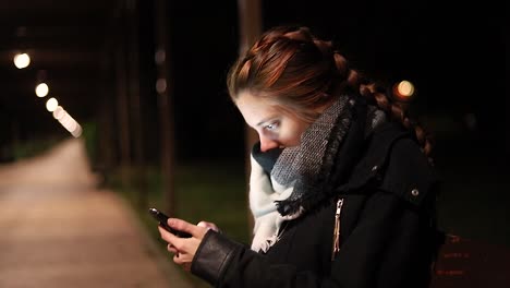 young woman looking their smart phone at night in a cold park bench