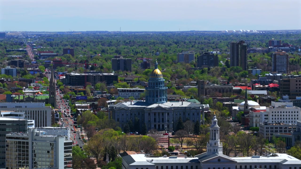 Luftfoto fra drone af Capitol Hill Civic Center Park i Downtown Denver, E. Colfax Avenue, en solrig forårsmorgen med blå himmel. Colorado Capitol Civic-bygningen og bygninger med gyldne kupler ses, med biltrafik på bygaderne, der drejer til højre.