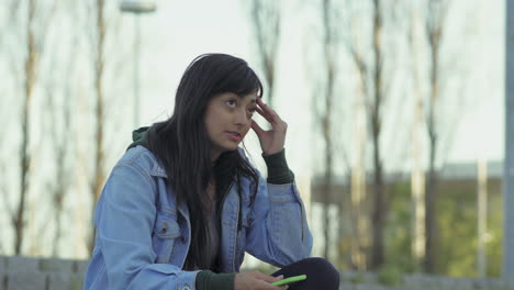 Thoughtful-teen-girl--holding-smartphone-while-sitting-outdoor.