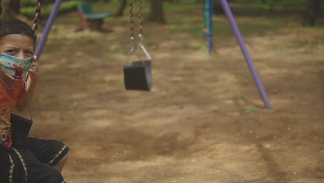 Asiatisches-Kind-Trägt-Gesichtsmaske-Und-Spielt-Auf-Der-Schaukel-Auf-Dem-Spielplatz,-Mittags,-Verspielter-Nachmittag-Auf-Dem-Spielplatz,-Goa