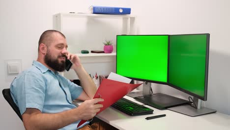 handsome freelancer man looking at folder, sitting against two monitors with green screen. worker speaks by mobile phone