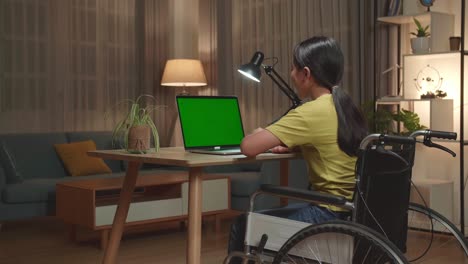 back view of asian kid girl sitting in a wheelchair while video call on laptop computer with mock up green screen at home