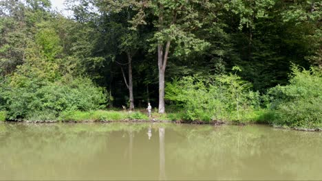 Male-angler-stands-on-the-shore-of-murky-lake-with-fishing-rod,-drone-wide-shot