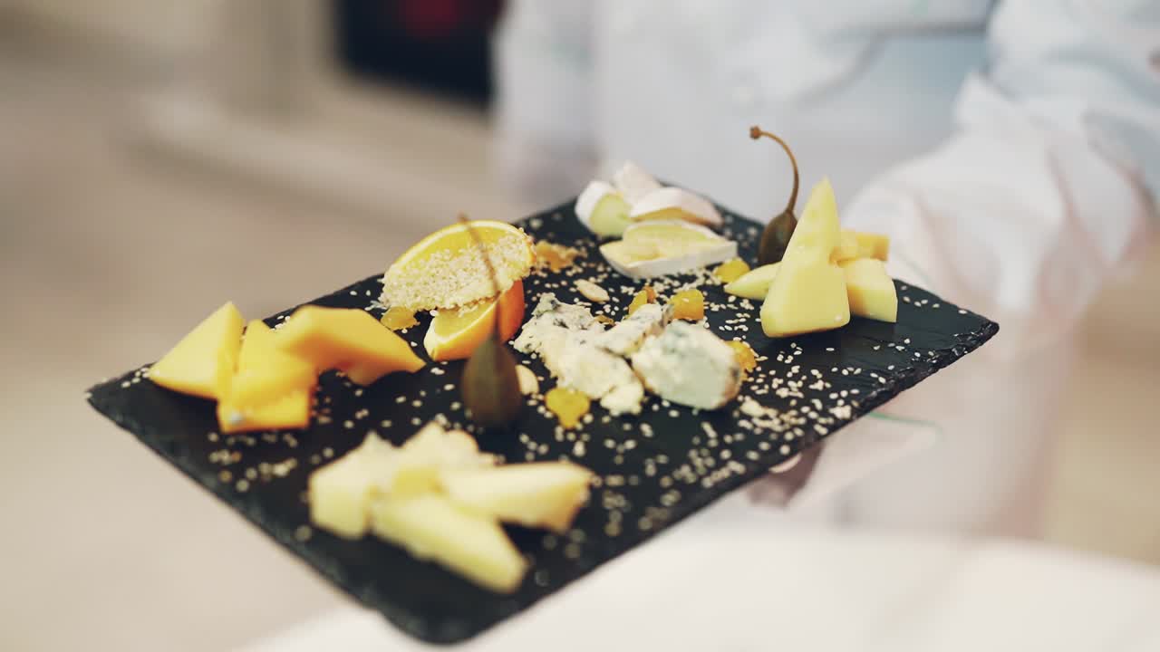 A waiter is holding in his hand a dark wooden board with different types of hard cheese decorated with fruit and sprinkled with sesame in the restaurant. Cheese tasting.