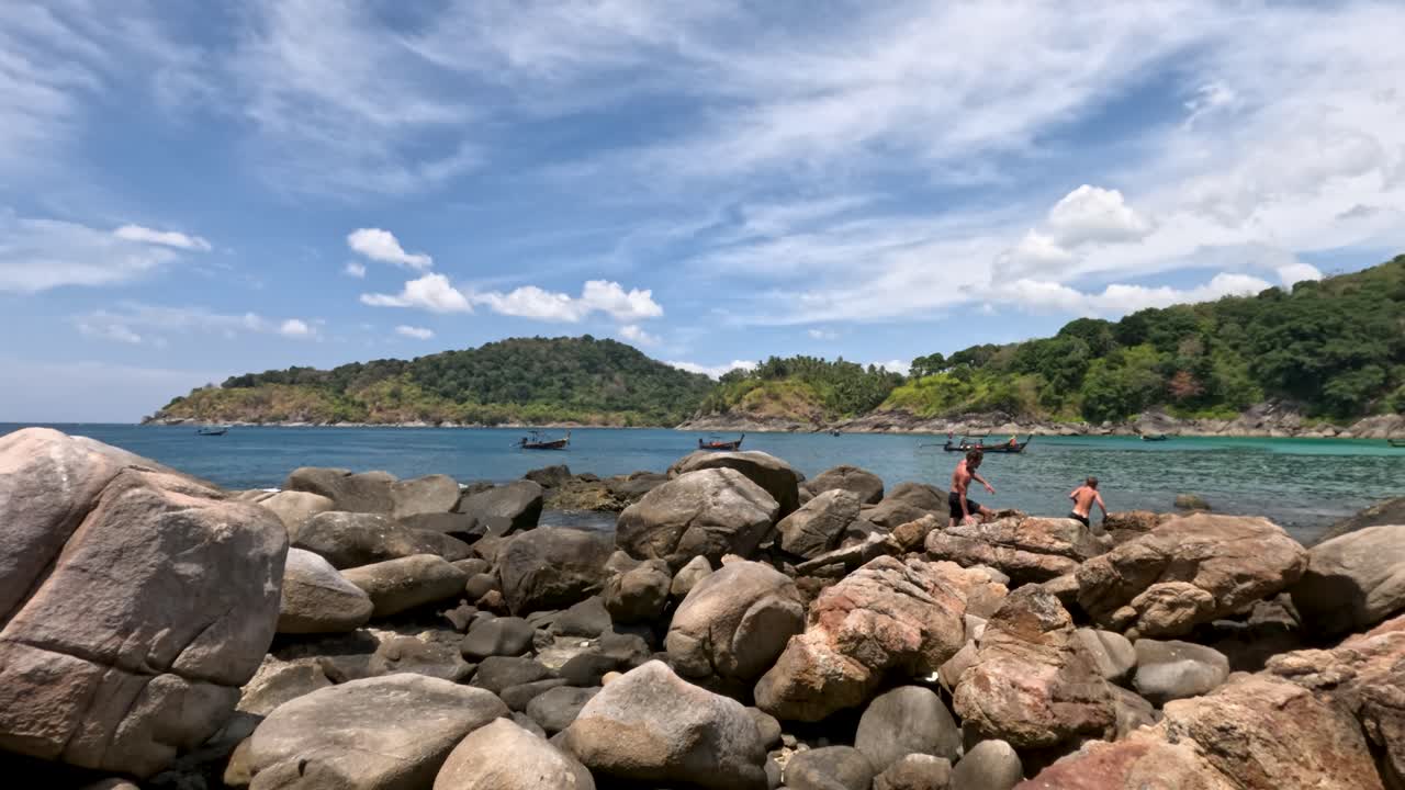 Two people climb large coastal boulders under bright daylight, tropical island and ocean background