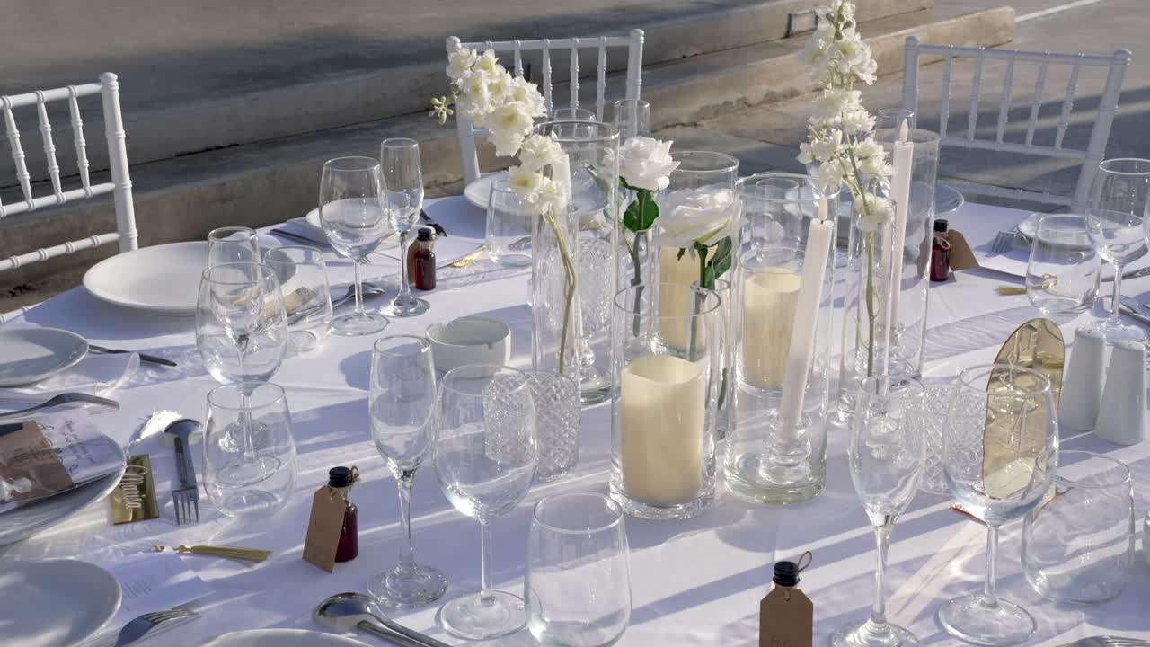 Et spisebord er dekket med glass, lys og blomster. Folk forbereder seg til en utendørs sammenkomst i et lyst og solrikt område. Bordet ser innbydende og organisert ut.