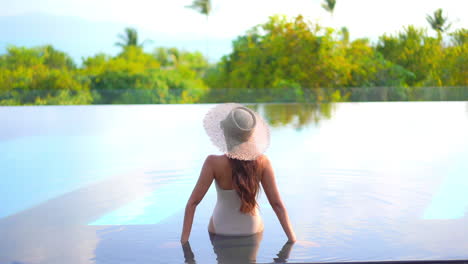 a woman with her back to the camera sits on the shallow step of a swimming pool as she looks out at the tropical landscape and ocean horizon beyond