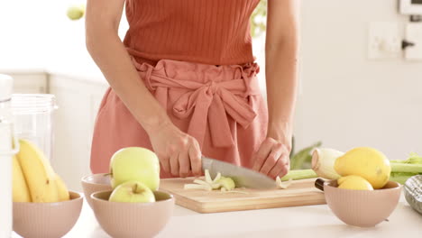 Woman chopping celery on wooden board in kitchen with fresh apples nearby