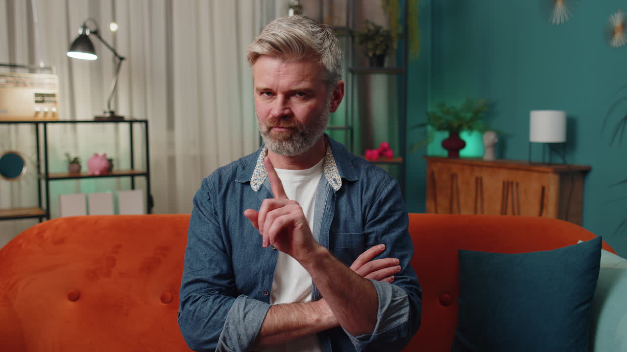 un hombre con barba y cabello gris se sienta en un sofá en una sala de estar y señala con un dedo a la cámara.