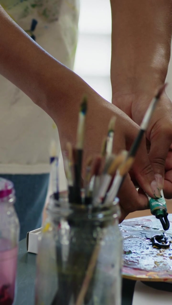 Vertical video: Opening cap, artist squeezing green paint onto palette to load brush, studio apron